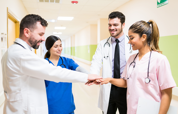Group of four clinicians in a hospital corridor putting hands together in a team huddle, smiling and collaborating.