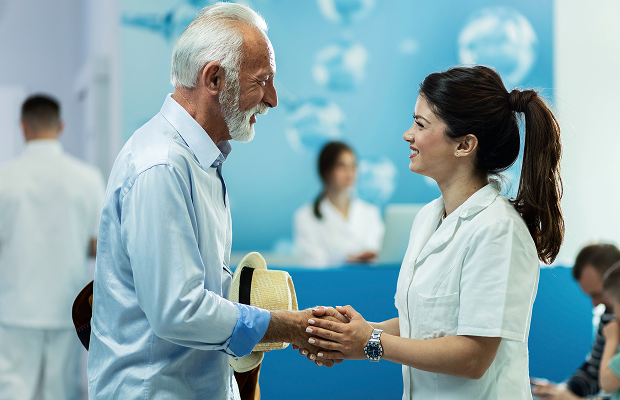 Elderly man shakes hands with a female healthcare professional in a clinic reception area, both smiling.
