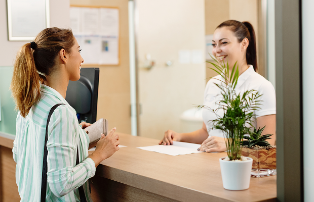 Guest with a striped shirt checks in at a front desk, smiling receptionist on the other side with a plant on the counter.