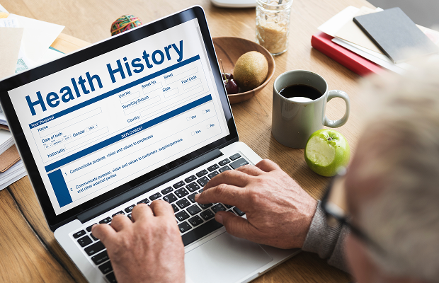 Person typing on a laptop displaying a health history form on a wooden desk. nearby are a mug, fruit, and papers.