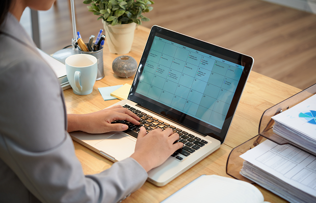 Person typing on a laptop at a desk with a calendar app open on the screen, surrounded by office supplies and a plant.
