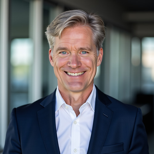 mike-emerson-mba-coo-axis-integrated-mental-health-denver-front-range-colorado Smiling man in a navy suit and white shirt, standing outdoors with blurred windows in the background.